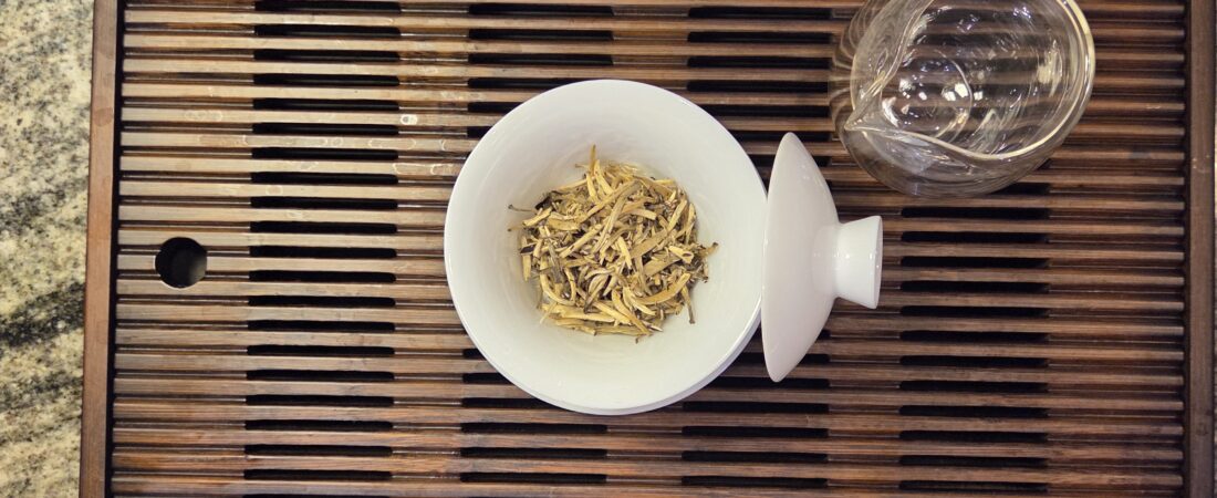 Dry whole tea leaves in an open white gaiwan beside a glass pitcher on a wooden tea tray.
