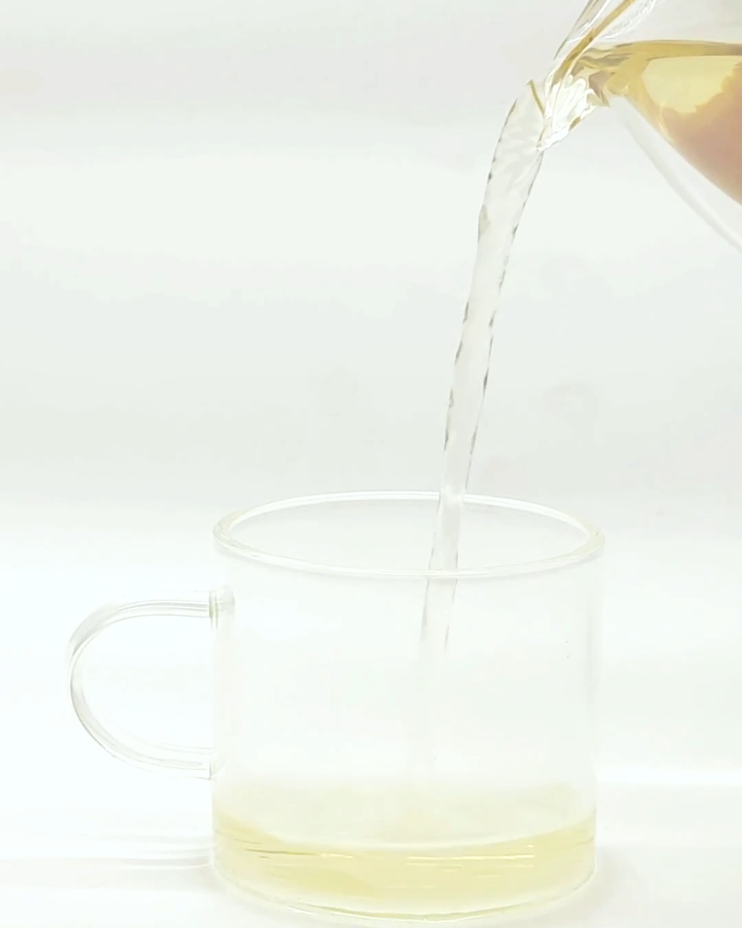 Pale tea poured from a glass pitcher into a clear handled cup against a white background.