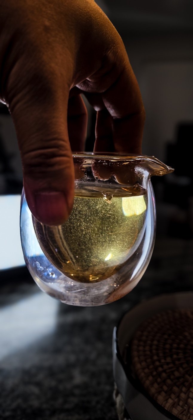 A hand holding a glass pitcher of golden tea against a dark background.